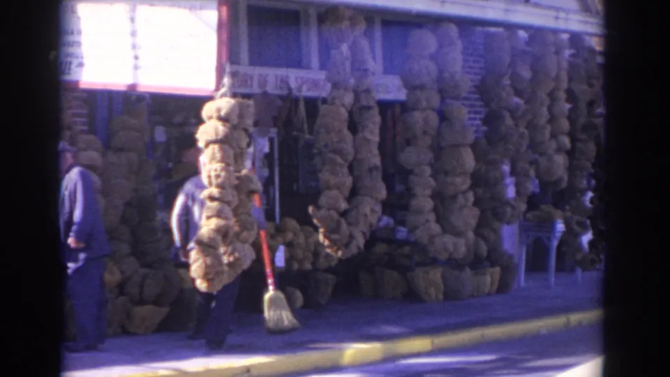 1962 Florida 8mm frame showing a man with a broom walking past a sponge shop display with a partial storefront sign.