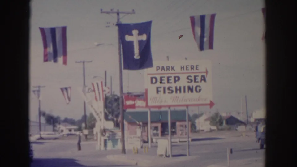 1962 Florida street-side view with a Deep Sea Fishing sign, striped flags, a cross banner, utility poles, and storefront frontage.