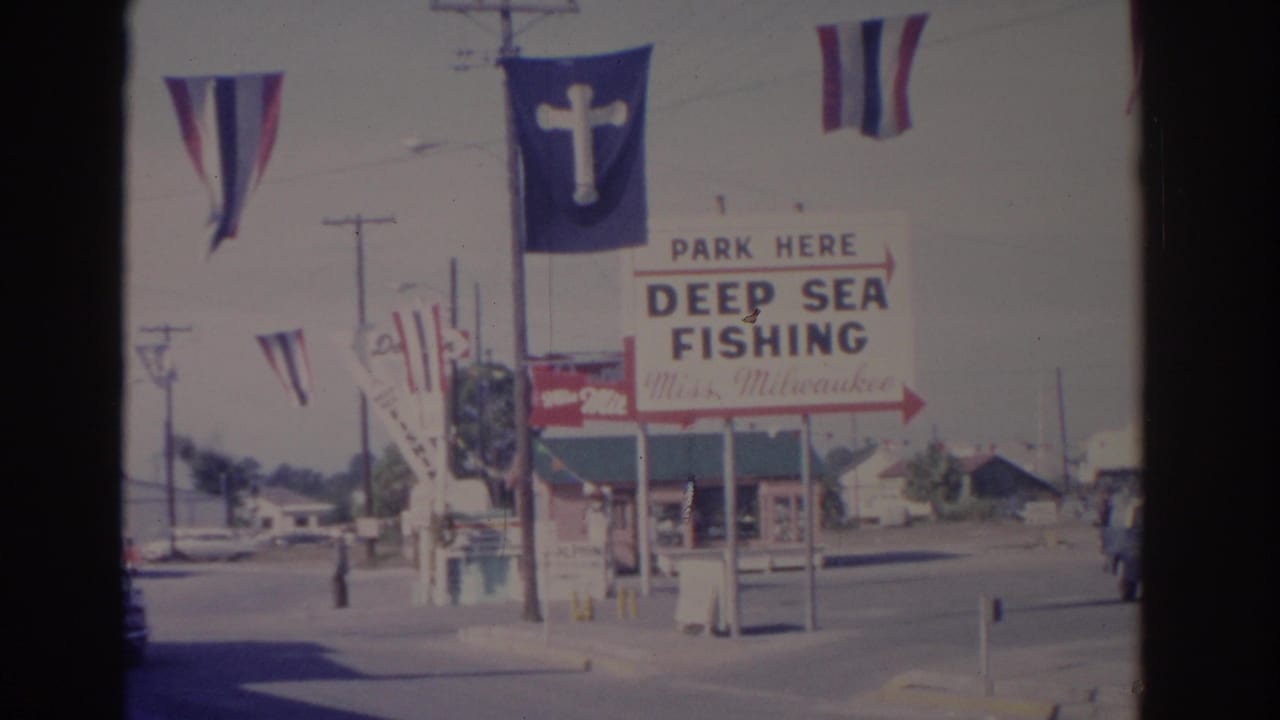 1962 Florida street view with a deep sea fishing sign, striped flags, a blue flag with a white cross, and roadside utility poles.