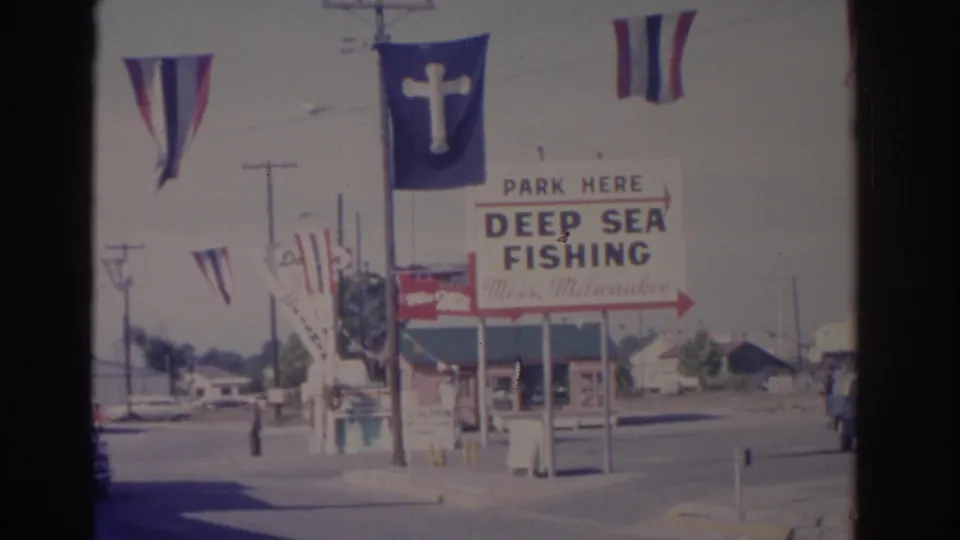 1962 Florida street view with a deep sea fishing sign, striped flags, a blue flag with a white cross, and roadside utility poles.