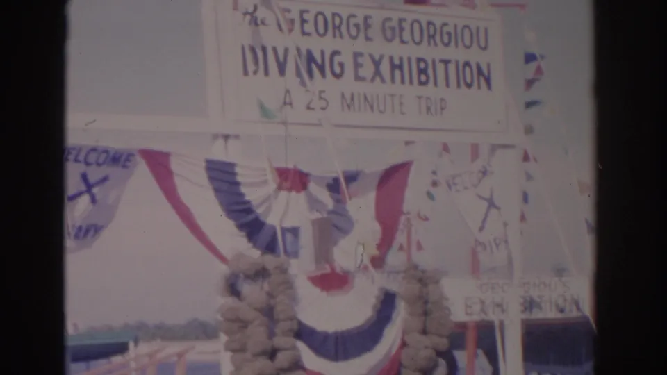 1962 8mm frame showing the George Georgiou Diving Exhibition sign, 25-minute trip text, bunting, flags, and entrance structure.