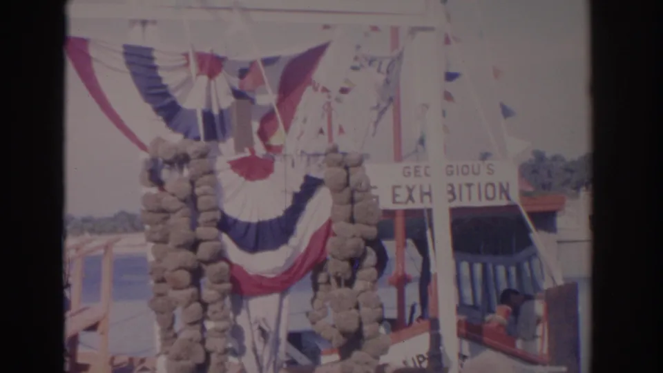 Florida tourist attraction entrance with patriotic bunting, exhibition signage, water in the background, and a small boat area with people nearby.
