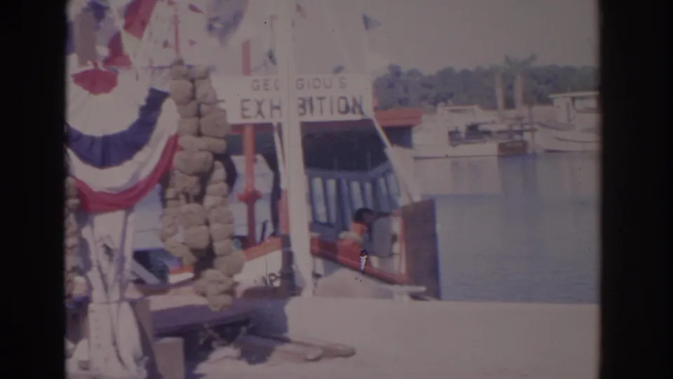 1962 Florida home-movie frame with bunting flapping near a docked vessel and small boats on calm harbor water.