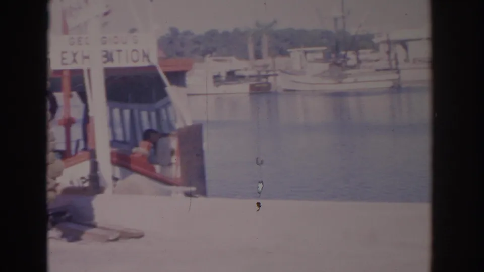 1962 Florida 8mm frame showing a decorated dockside vessel, calm harbor water, and a seated person partly visible near the boat.
