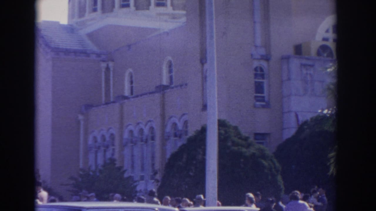 Crowd outside a light-colored arched building with a pole and parked car in front.