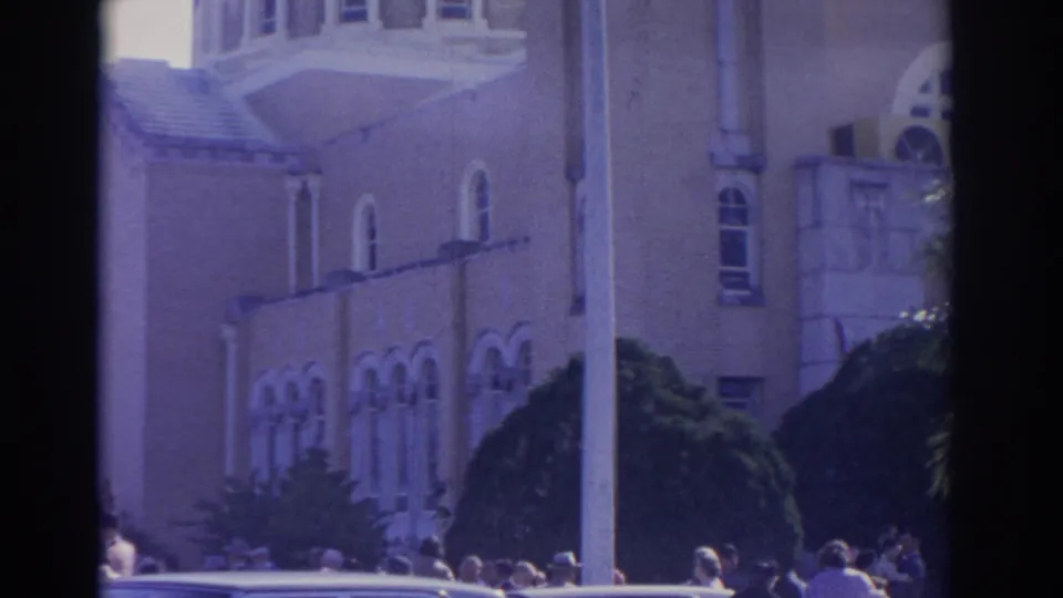 1962 Florida street view of people gathered outside a light-colored building with arched windows, a pole, shrubs, and cars at the curb.