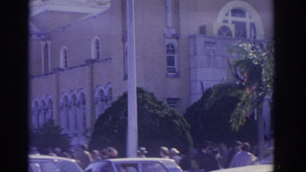 Crowd and cars in front of a light-colored building with arched windows and shrubs.