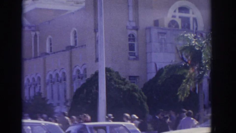 1962 Florida street scene with a crowd near a formal building, visible arched windows, trimmed shrubs, palm foliage, and mid-century cars.