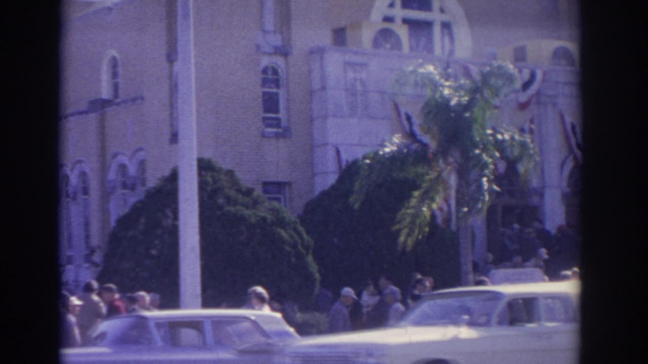 1962 Florida home-movie frame showing a palm tree, people gathered by a decorated building facade, and light-colored early-1960s cars.