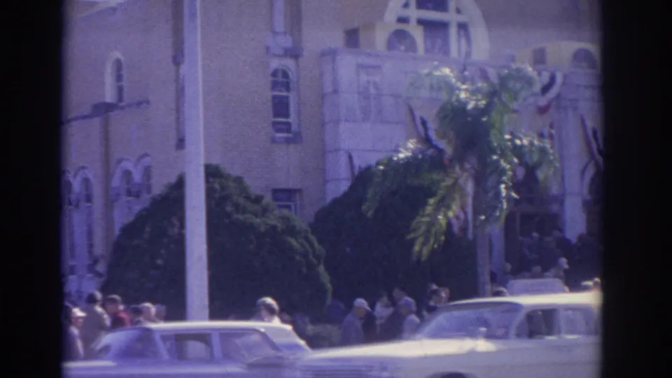 1962 Florida home-movie frame showing a palm tree, people gathered by a decorated building facade, and light-colored early-1960s cars.