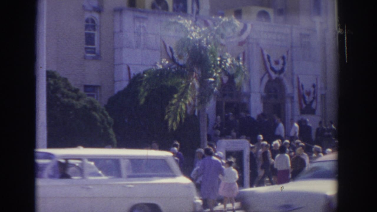1962 Florida street view with people gathered near a decorated building entrance, palm foliage, a light station wagon, and sunlit cars.