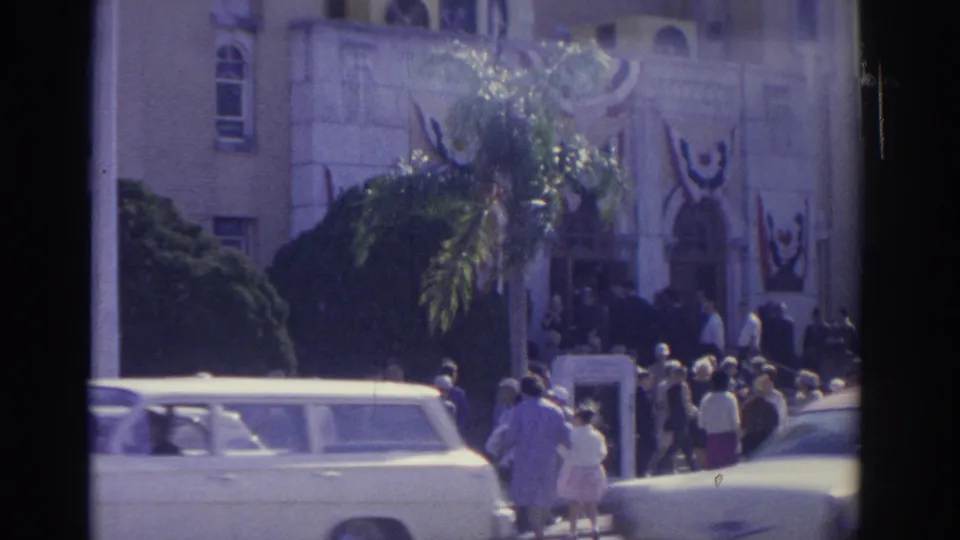 1962 Florida street view with people gathered near a decorated building entrance, palm foliage, a light station wagon, and sunlit cars.