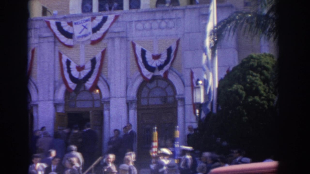 1962 Florida home-movie frame showing a decorated public building facade with bunting, spectators, and people near the entrance.