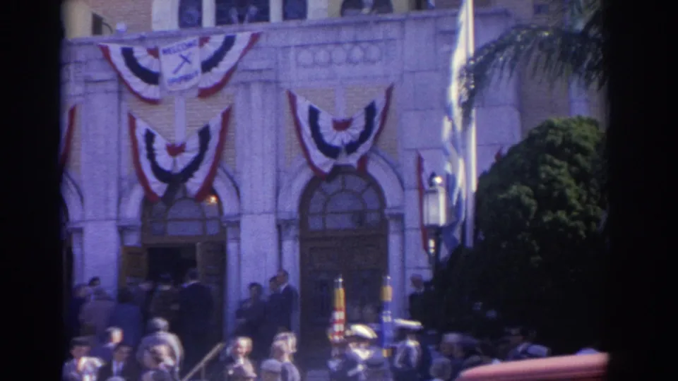 1962 Florida home-movie frame showing a decorated public building facade with bunting, spectators, and people near the entrance.
