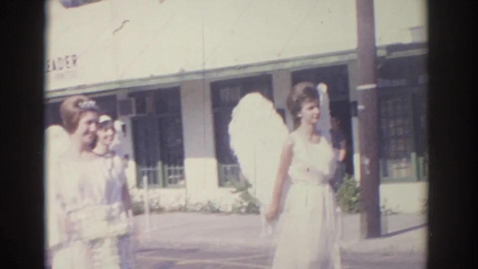 Women in white dresses with large feathered angel wings stand on a sunny Florida street near storefront windows and signage.