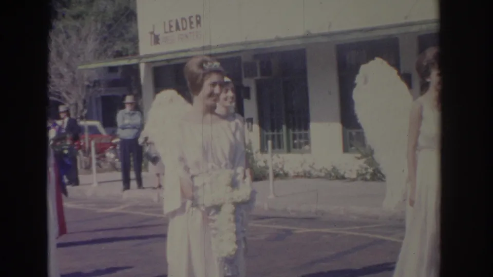 A woman in a white angel costume with feathered wings holds a white bouquet in a 1962 Florida street-event scene.