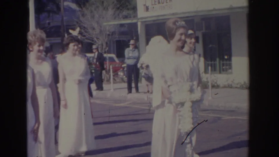 Women in white angel-style costumes pass a storefront with visible signage while onlookers stand along the sunny Florida street.