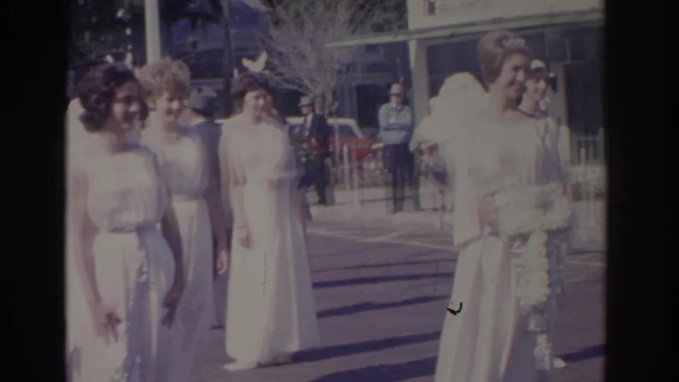 1962 Florida home-movie frame of pageant contestants in white gowns and angel wings standing downtown with bystanders behind them.