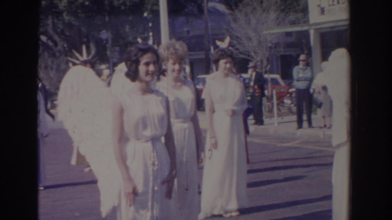 Contestants in angel wings stand in formation near storefronts and parked cars.