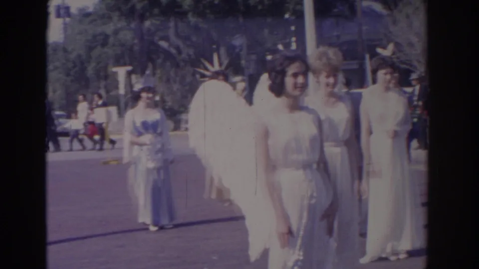 1962 Florida home-movie view of women in white gowns and angel wings waiting along a downtown street with bystanders behind them.