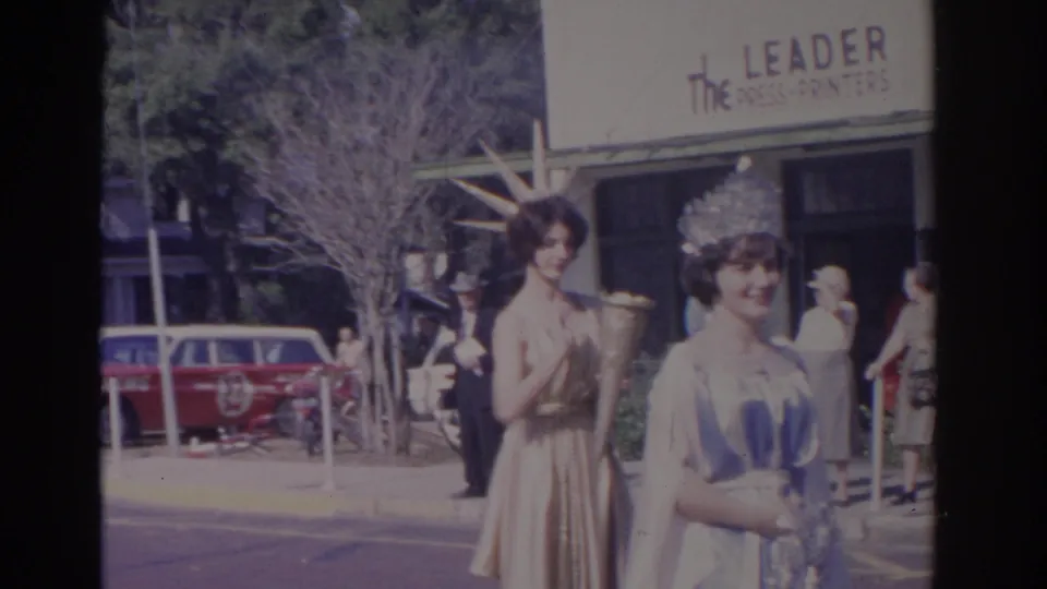 1962 Florida frame showing costumed pageant participants near a readable The Leader Press-Printers sign and parked cars downtown.