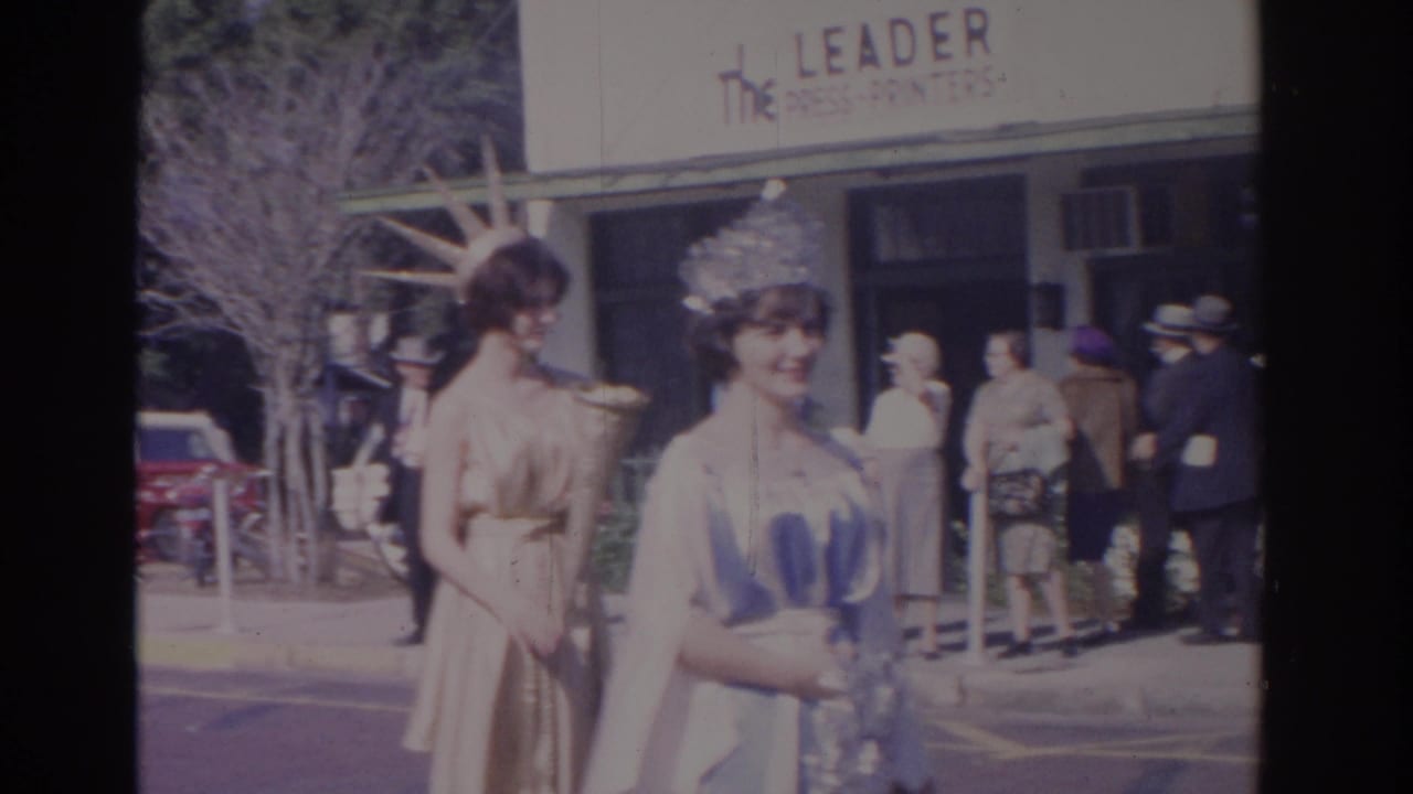 Two young women in crowns and gowns pass spectators outside The Leader Press-Printers storefront.