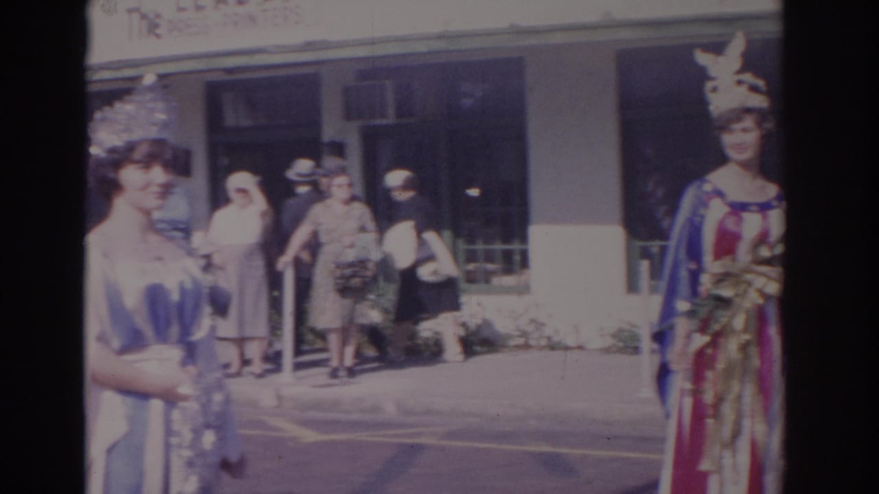 A crowned woman in a blue and white gown stands near another costumed woman and sidewalk spectators.