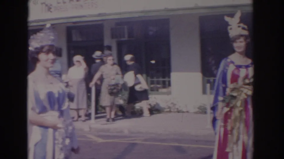 1962 Florida parade frame with a crowned woman in a blue and white gown near a patriotic-costumed woman and spectators outside a storefront.