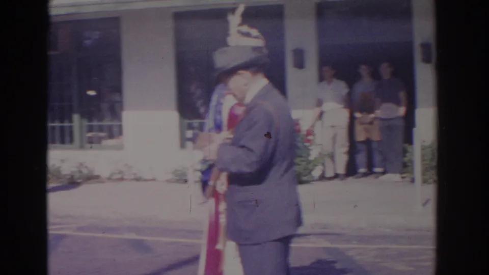 1962 Florida 8mm frame showing a man in a grey suit and hat beside a woman in a patriotic costume, with spectators behind them.