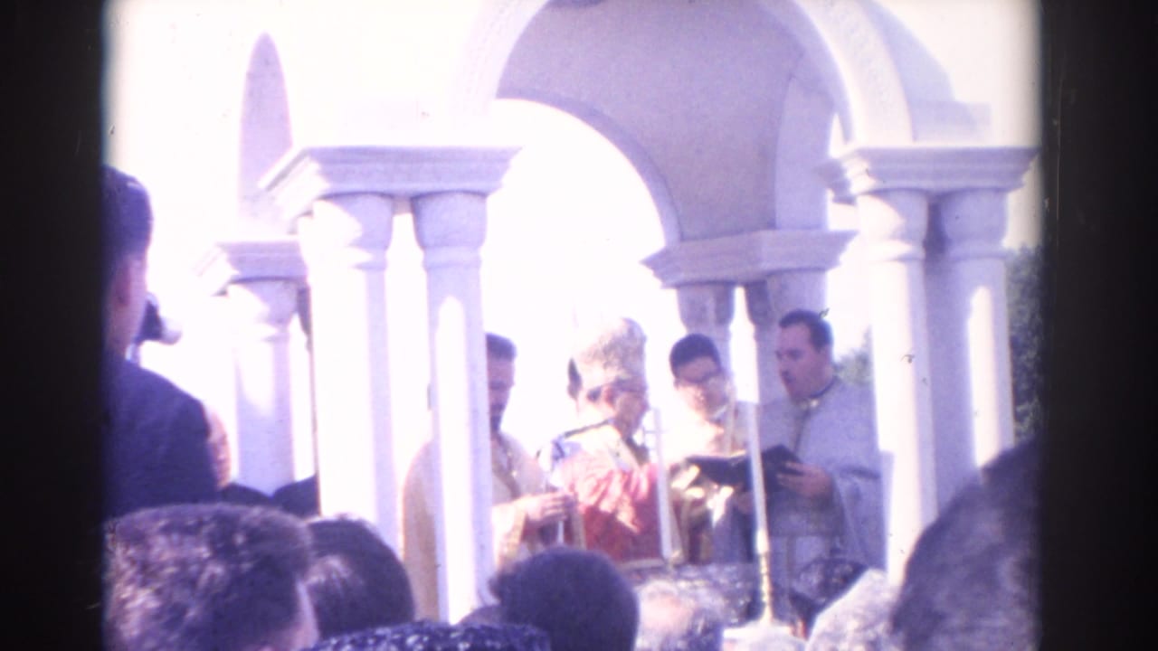 1962 Florida home-movie frame showing clergy at an outdoor Catholic mass beneath white arches, partly blocked by the crowd.