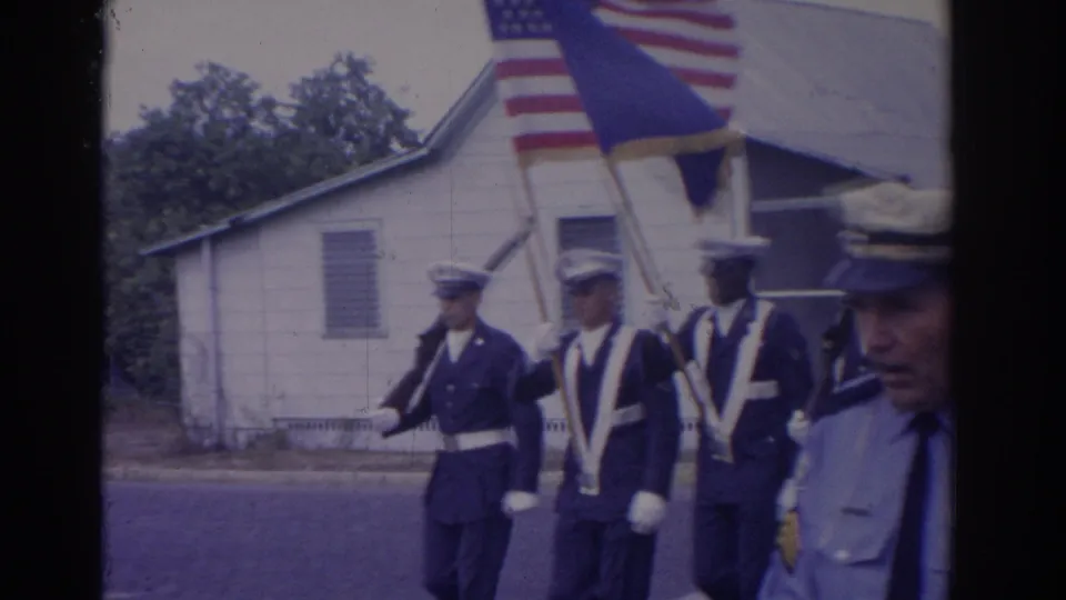 1962 Florida home-movie frame showing uniformed marchers in a street parade with an American flag and blue banner.