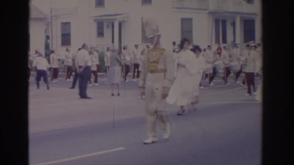 1962 Florida parade view with a cream-uniformed marcher, women in white dresses, and band members moving along a residential street.