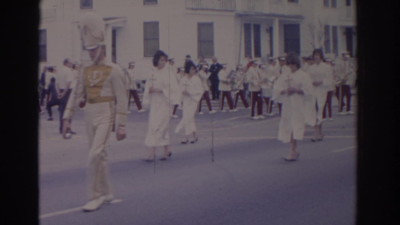 Curbside 8mm view of a Florida parade with a cream-uniformed marcher, cross-bearing women, and a marching band behind them.
