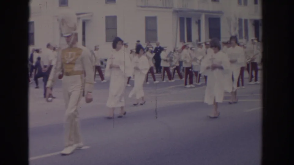 Curbside 8mm view of a Florida parade with a cream-uniformed marcher, cross-bearing women, and a marching band behind them.