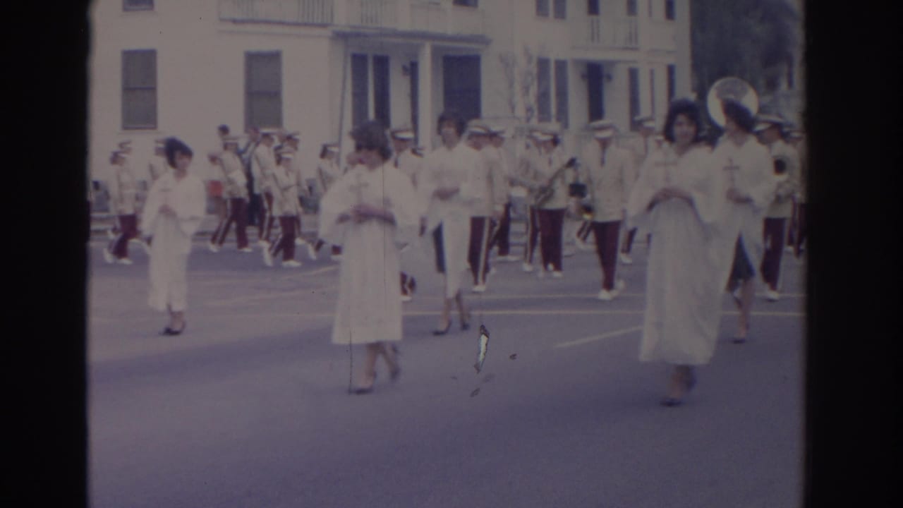 1962 Florida parade frame showing women in white dresses with crosses, followed by band members in light uniforms on a city street.