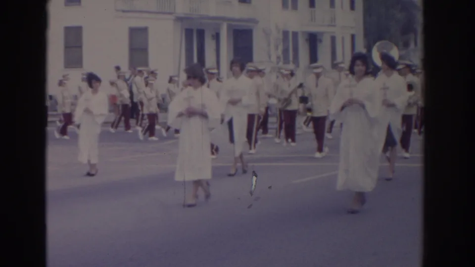1962 Florida parade frame showing women in white dresses with crosses, followed by band members in light uniforms on a city street.