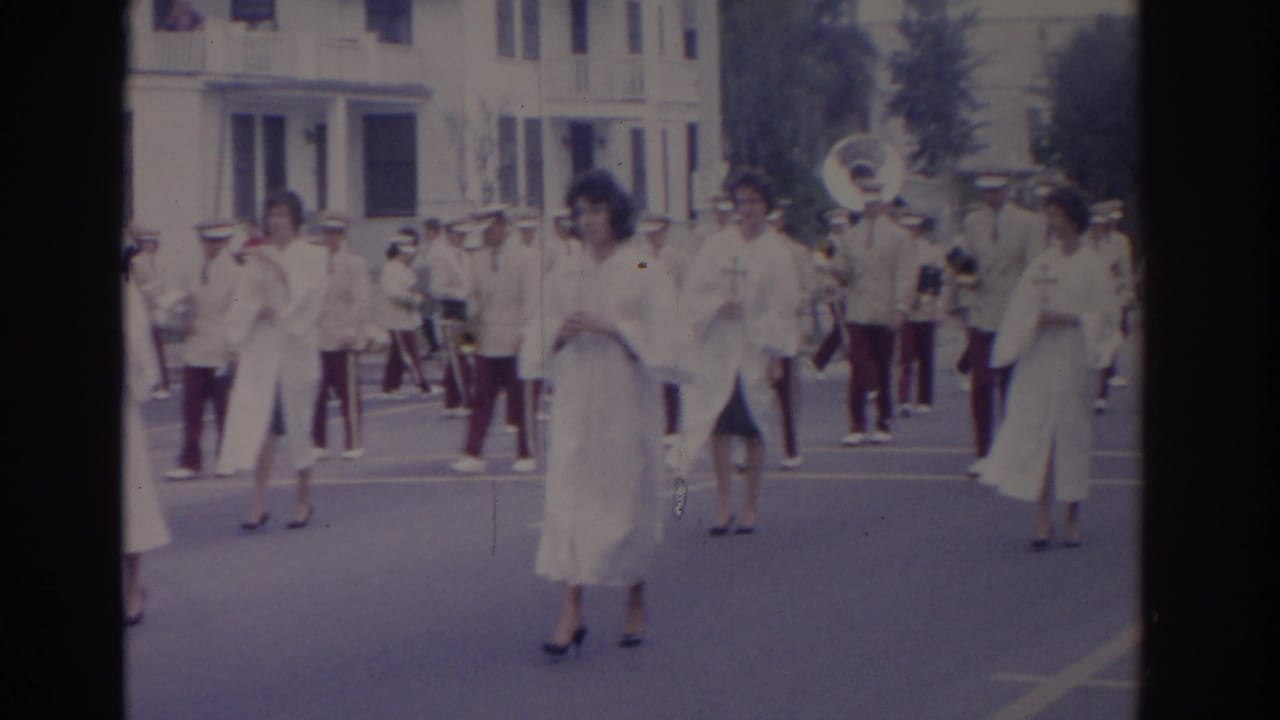 Florida 1962 home-movie frame with women in white dresses, a visible sousaphone, and marching band members moving along the street.