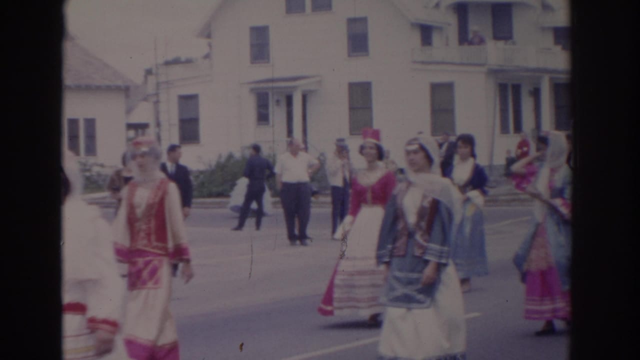 In 1962 Florida home-movie footage, women in colorful historical-style dresses walk in a street parade past onlookers and white clapboard houses.
