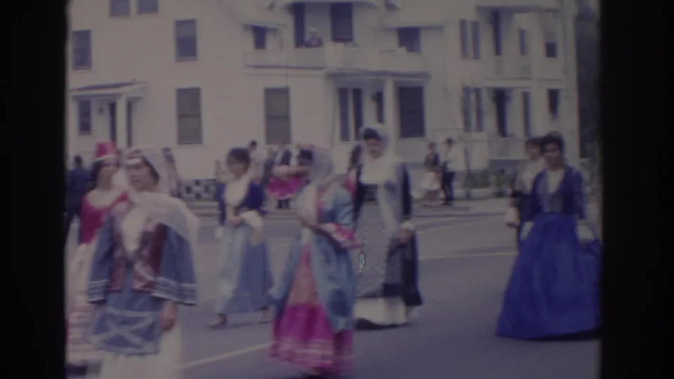 Costumed marchers in long dresses and headscarves proceed along a Florida residential street in 1962, with spectators and houses in the background.