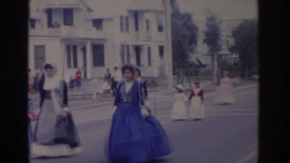 A woman in a blue historical-style dress passes near the camera during the 1962 Florida parade, with other marchers and spectators farther down the street.