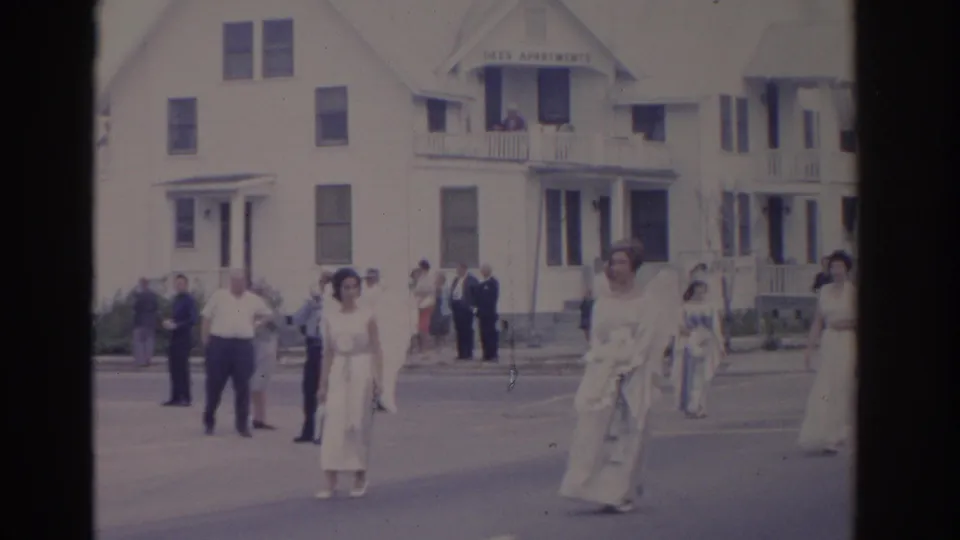 Women in pale period-style dresses walk through a Florida residential street parade in 1962, with spectators gathered near white apartment buildings.