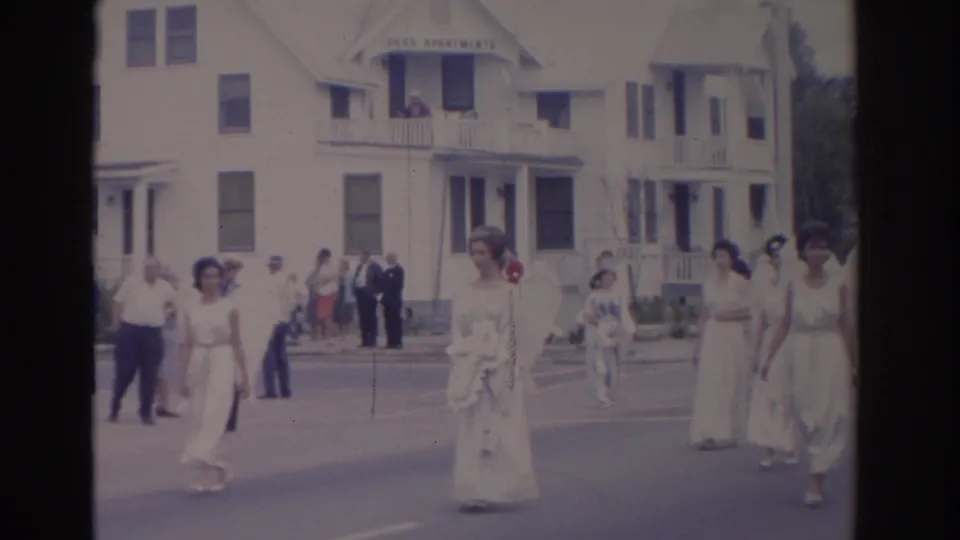1962 Florida home-movie frame showing women in white gowns and angel-wing costumes walking past spectators near Dees Apartments.