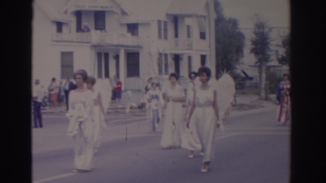 Parade participants in white dresses walk in the street with onlookers behind them.