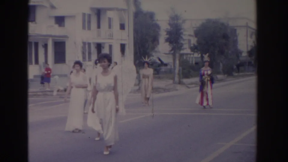 Two women in white dresses approach the camera in a 1962 Florida street parade, with a colorful costumed figure farther back.