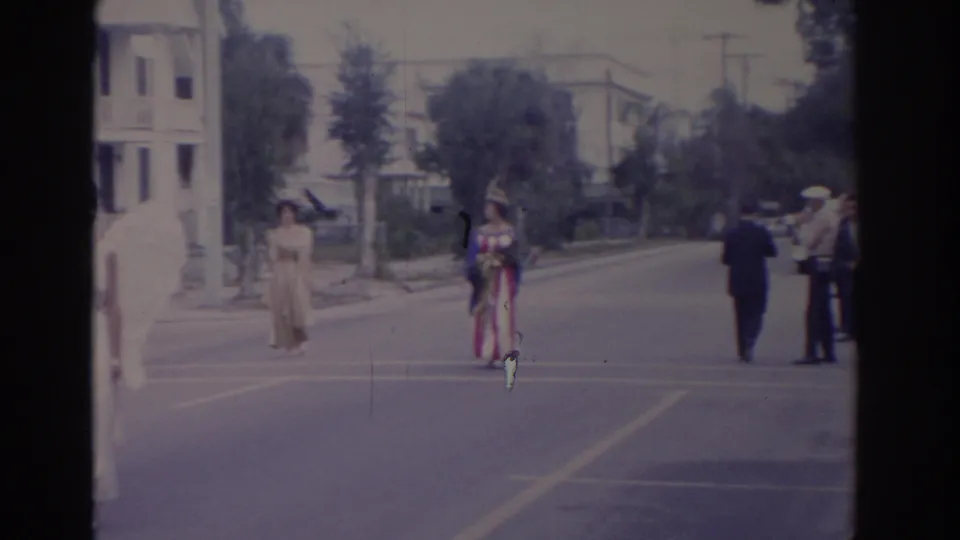 Later frame from the 1962 Florida parade showing a colorful costumed participant, spectators, and a residential street setting.