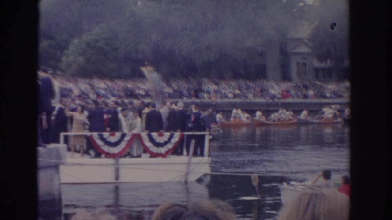 Decorated boat with bunting passes a crowded riverbank during a public waterfront event.
