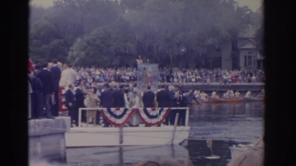 A 1962 Florida river-parade view with spectators packed along the bank and a bunting-trimmed boat carrying adults in formal clothing.