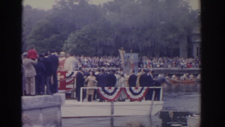 Home-movie view from a Florida waterfront event, with a decorated boat in the foreground and dense spectators across the river.