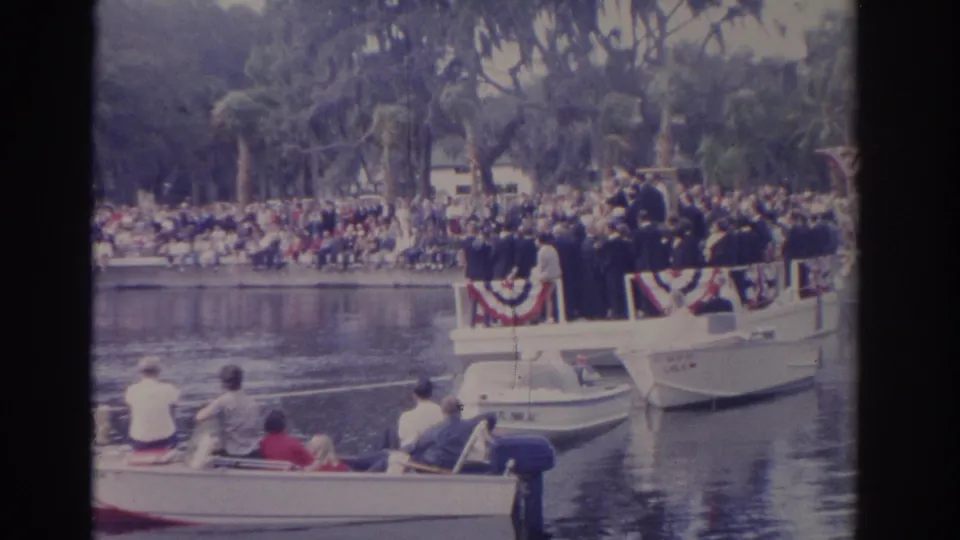 1962 Florida river parade frame showing a bunting-trimmed boat with formal passengers, nearby small boats, and spectators along the shore.