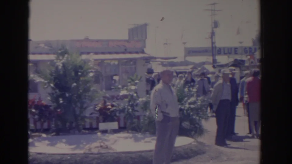 1962 Florida fairground frame showing men near landscaped displays and booths, with a partial blue sign in the background.
